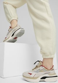 PUMA-Sneaker haben ein beiges Obermaterial mit roten Akzenten und einem strukturierten grauen Paneel. Die Sohle ist klobig mit einem schwarzen Abschnitt und einem profilierten Muster.