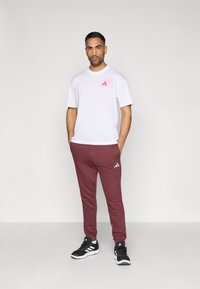 Biele tričko s krátkym rukávom a ružovým logom, doplnené bordovými teplákmi so strieborným logom; model má na sebe čierne športové topánky.