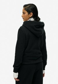 Felpa nera con cappuccio dalla vestibilità comoda, caratterizzata da un ampio cappuccio, polsini a costine e una texture morbida. Il modello indossa sotto delle maniche bianche a strati.