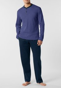 Uomo in maglietta a maniche lunghe di colore viola e pantaloni blu scuro a vestibilità comoda, in piedi a piedi nudi su uno sfondo neutro.