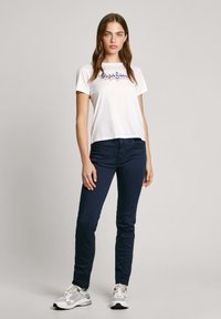 Bijela majica kratkih rukava s plavim i crvenim "Pepe Jeans" logotipom, u kombinaciji s tamnoplavim uskim hlačama. Model nosi ležerne tenisice.