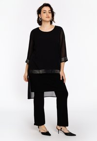 Schwarze Chiffon-Tunika mit Dreiviertelärmeln, verziertem Saum, die über schwarzen enganliegenden Hosen getragen und mit spitzen schwarzen Absätzen kombiniert wird.