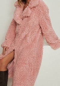 Abrigo de piel sintética rosa con una superficie texturada y esponjosa; cuenta con un gran cuello y mangas largas, ofreciendo un ajuste oversize y una apariencia cálida.