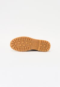 Tan rubber sole with a textured grip pattern, featuring hexagonal lugs and a rectangular logo in the center. White background.