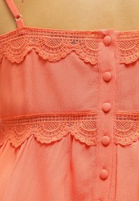 Persikofärgad blus med spetsdetaljer vid halsringningen och ryggen, med sex tygklädda knappar i mitten och en lätt textur.