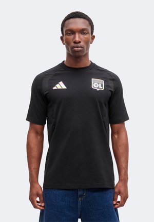 Mężczyzna w czarnej koszulce klubu piłkarskiego Olympique Lyonnais z logo Adidas, stojący na jednolitym jasnym tle.