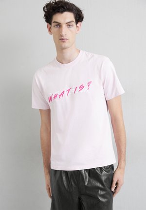 Light pink cotton t-shirt with short sleeves, featuring bold pink text "WHAT IS?" diagonally and a relaxed fit. Worn with black shiny trousers.