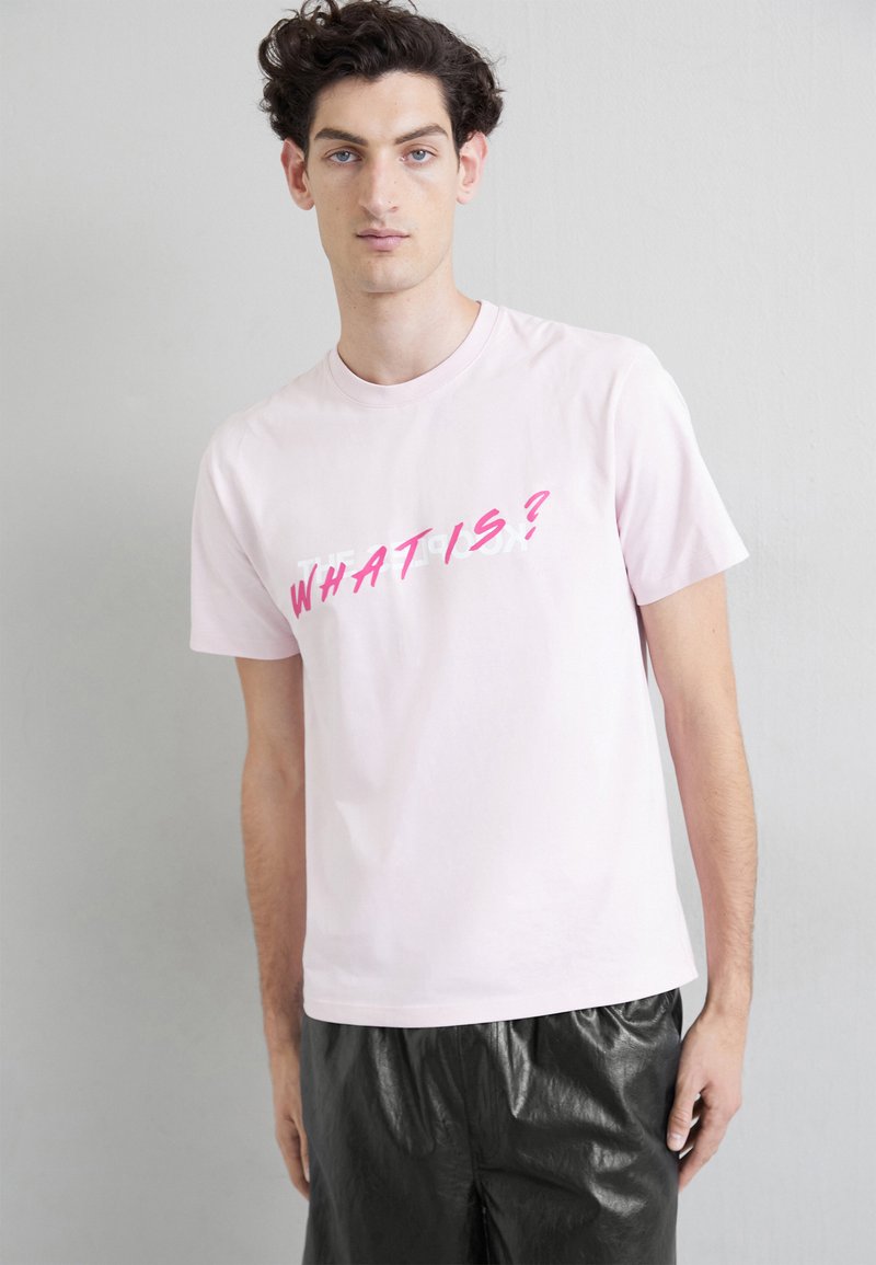 Lys rosa bomulls-t-skjorte med korte ermer, med fet rosa tekst "HVAD ER?" diagonalt og en avslappet passform. Worn med svarte, blanke bukser.
