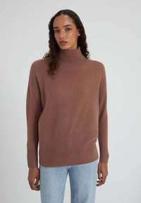 Frau mit lockigem Haar, die einen braunen Rollkragenpullover und hellblaue Jeans trägt, steht vor einem schlichten hellen Hintergrund.