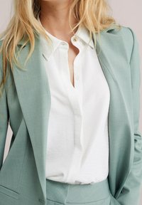 Blazer verde claro hecho de tela texturizada con una camisa blanca lisa debajo. La camisa tiene cuello y una tapeta con botones.