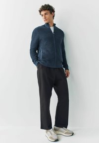 Fiatal férfi áll egy fehér falnak támaszkodva, sötétkék cipzáras pulóvert, fekete nadrágot és bézs sportcipőt visel, amelynek fehér fűzője van.