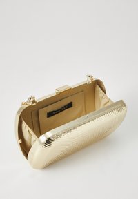 Gold metallic clutch with wave-patterned texture, rectangular shape, and a secure clasp. Interior features a beige lining and brand label.