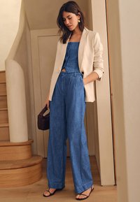 Smėlio spalvos švarkelis dėvimas virš mėlynos džinsinės trumpintų marškinėlių ir plačių kelnių. Juodos suvarstomos basutės ir mažas rudas rankinukas. Lygios tekstūros ir švarios linijos.