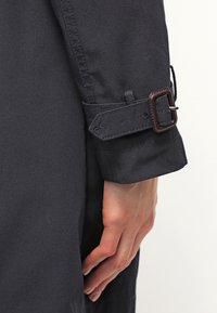 Gros plan sur la main d'une personne reposant le long de son côté, portant une manche de manteau sombre avec une sangle réglable et une boucle carrée.