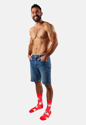 Chaussettes rouges avec des motifs de carlin, shorts en denim, torse nu et teint clair, sur un fond blanc uni.