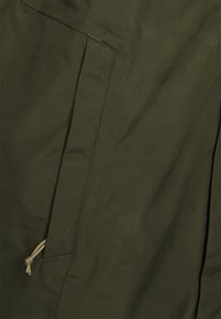 Materiał oliwkowozielony o gładkiej fakturze, z boczną kieszenią i detalem w formie sznureczka, podkreślający minimalistyczny design.