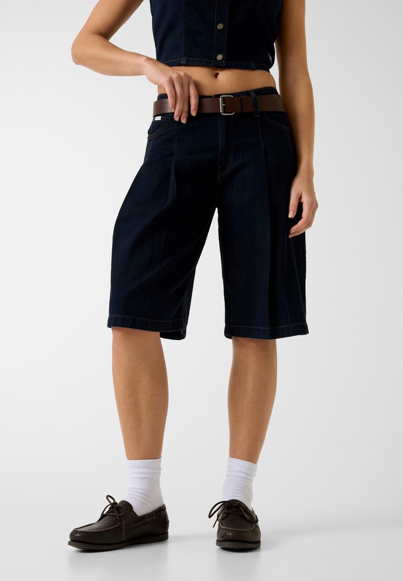 Jeune femme portant un short bermuda en denim bleu foncé avec une ceinture marron, des chaussettes blanches et des mocassins marron foncé, debout devant un fond uni.