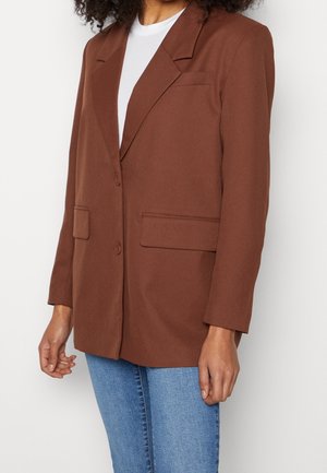 Femme portant un blazer marron sur une chemise blanche et un jean bleu, debout devant un fond clair uni.