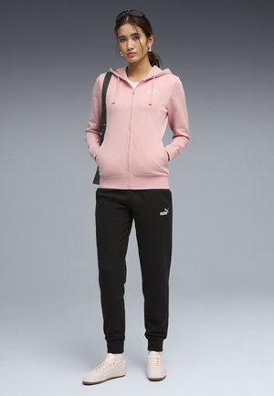Femme debout portant un sweat à capuche zippé rose, un pantalon de survêtement noir Puma, des baskets rose clair, des chaussettes blanches et des lunettes de soleil sur la tête.