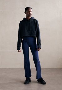 Svart cropped hettegenser med snortrekk, kombinert med høy midje mørkeblå vide bukser. Modell iført svarte treningssko, står på en lys overflate.
