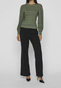 Olivegrünes, strukturiertes Langarm-Oberteil mit Rundhalsausschnitt und schwarzen, taillierten Hosen. Das Model trägt schwarze, spitze Pumps.