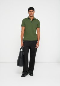 Oliven grønn polo skjorte med krage, paret med svarte vide bukser. Modellen holder en svart lær totebag, stående mot en hvit bakgrunn.