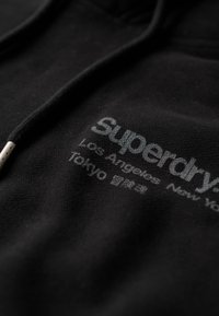 Sudadera negra con un acabado texturizado que presenta un logo impreso. Incluye costuras decorativas y cordones con puntas de metal.