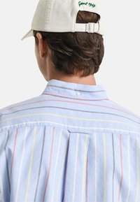 Camisa a rayas azul claro con rayas verticales en rojo, amarillo y gris, con un detalle de cuello abotonado en la parte posterior y un corte casual.