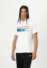 Biela bavlnená tričko s farebnou grafikou vĺn, na pozadí s ružovým a modrým gradientom a bielym logom Nike v strede.