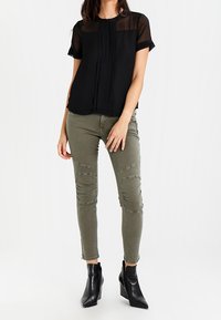 Černá blůza s krátkým rukávem z průsvitné tkaniny a s plisováním, kombinovaná s úzkými olivově zelenými džíny s detaily švů na kolenou a po stranách. Černé kotníkové boty.