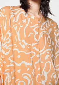 Blusa arancione con motivo astratto a spirale bianca, scollo rotondo e dettaglio di bottoni frontali. Realizzata in tessuto leggero con una texture liscia.