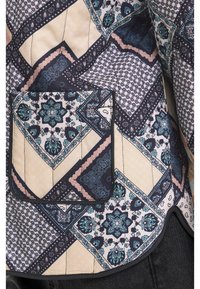 Quiltet jakke med et geometrisk mønster i marineblå, creme og grønblå. Har en stor lomme med lap og karakteristiske teksturerede stofdesigns.