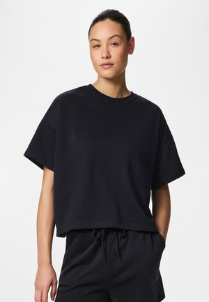 Fiatal nő hátrasimított hajjal, laza fekete rövid ujjú pólót és hozzáillő, fekete, zsinórral állítható rövidnadrágot viselve, fehér háttér előtt.