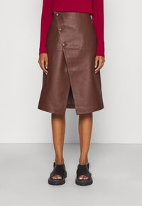 Ruskea nahkainen kääntöhame, polvenpituinen malli, jossa edessä halkio, suuret napit ja sileä pinta. Yhdistetty mustiin sandaaleihin.