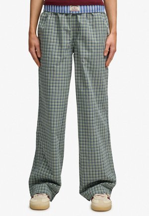 Person iført løse grønne og blå ternede bukser med en blåstribet linning og beige sneakers, stående med hænder afslappet ved siden.