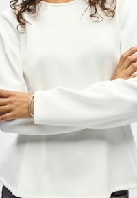 Haut à manches longues blanc avec un col rond et un ourlet évasé. Texture douce avec une finition lisse. Le bras est orné d'un accent de bracelet en argent.