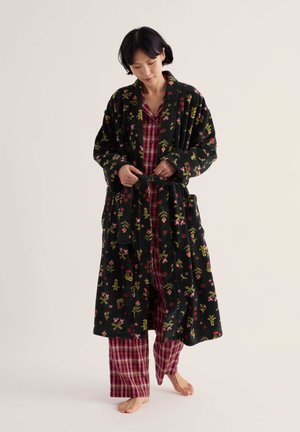Femme portant un pyjama à carreaux rouges attachant la ceinture d'un peignoir noir à motif floral, debout pieds nus sur un fond uni.