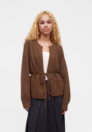 Jeune femme aux cheveux blonds bouclés portant un cardigan marron duveteux noué à la taille sur un haut blanc et une jupe plissée foncée, debout devant un fond uni.