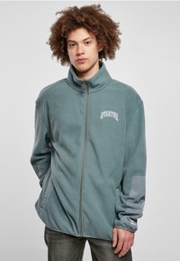 Fleece-tyylinen vetoketjullinen takki merensinisenä, harmailla yksityiskohdilla. Ominaisuuksiin kuuluu korkea kaulus ja sivutaskut, kevyt rakenne sekä brodeerattu "STARTER"-logo.