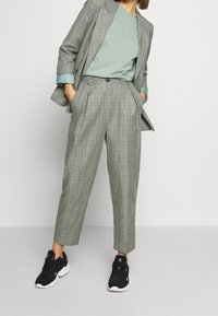 Woman wearing gray plaid suit with cropped pants and jacket over a light green shirt, paired with black sneakers, standing hands in pockets.