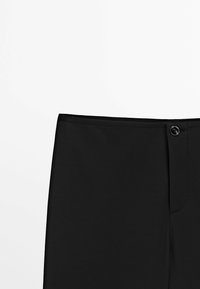 Partie supérieure d'un pantalon noir avec une taille à bouton et une couture visible sur fond blanc.