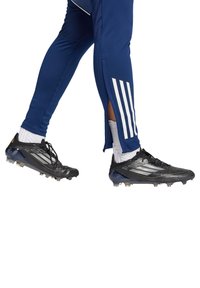 Schwarze Fußballschuhe mit weißen Akzenten, kombiniert mit marineblauen Sporthosen, die drei weiße Streifen und ein seitliches Schlitzdesign aufweisen.
