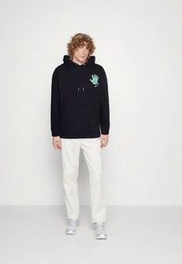 Black hoodie with an embroidered mint green hand logo on the left chest, paired with white pants and gray sneakers. Casual design.