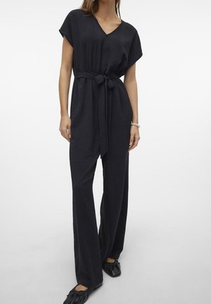 Vrouw in een zwarte V-hals jumpsuit met een gestrikt tailleband en zwarte ballerina's, staand tegen een effen witte achtergrond.