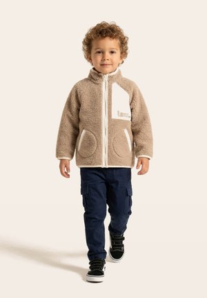 Young child walking forward wearing beige fleece jacket, navy cargo pants, and black sneakers with white soles.