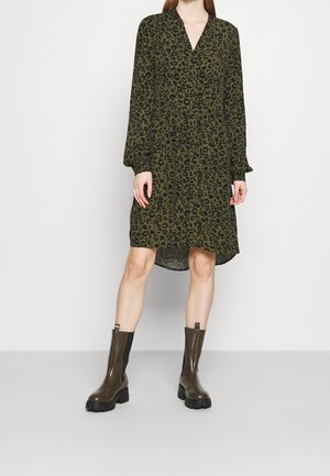 Femme portant une robe à manches longues imprimé léopard olive et noir avec col en V, associée à des bottines épaisses foncées, debout sur fond blanc.