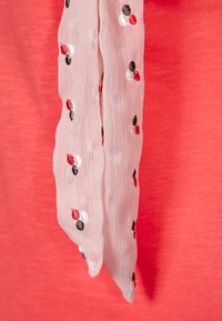 Jasnopinkowy szalik z haftowanymi kropkami w kolorze czerwonym, czarnym i białym, ułożony na koralowym tle, o miękkiej i lekkiej teksturze.