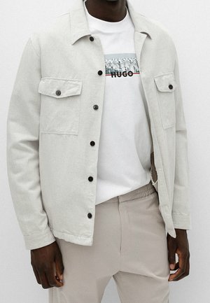 Uomo con giacca beige abbottonata sopra una T-shirt bianca con logo "HUGO" nero e rosso e pantaloni beige su sfondo semplice.