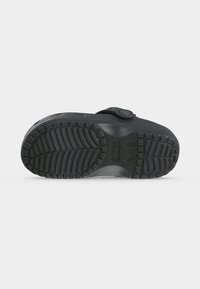 Sabot Crocs noir montré par la semelle, avec une semelle texturée pour l’adhérence et une bride arrière avec détail bouton.