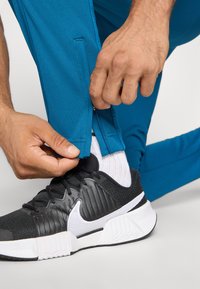 Rankos užsegia blauzdos užtrauktuką ant mėlynų kelnių, uždėtų ant baltų kojinių ir juodų "Nike" sportbačių su balta soliuku, priešais vientisą fono.